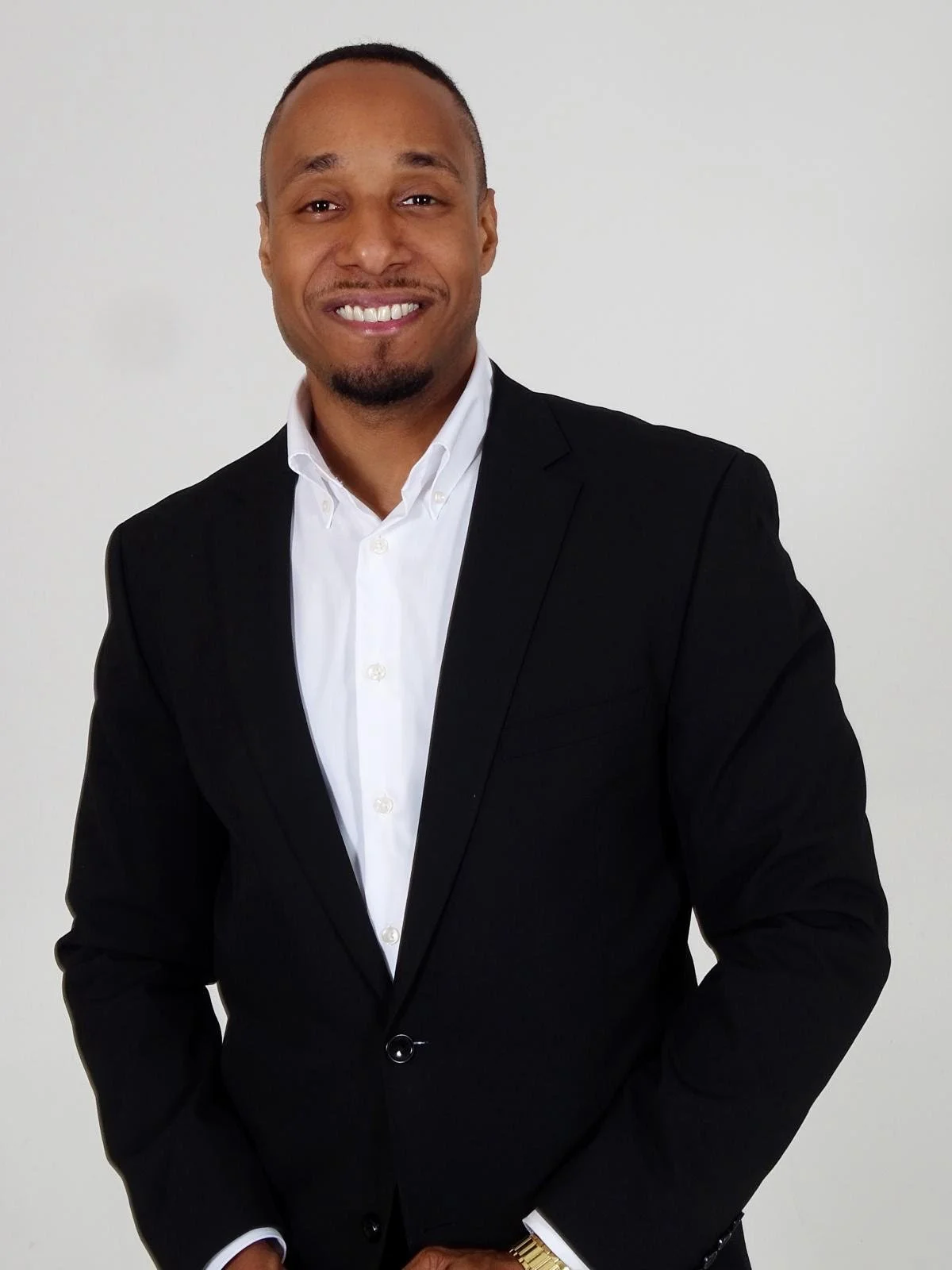Professional man wearing a black suit and white shirt, smiling against a plain white background.