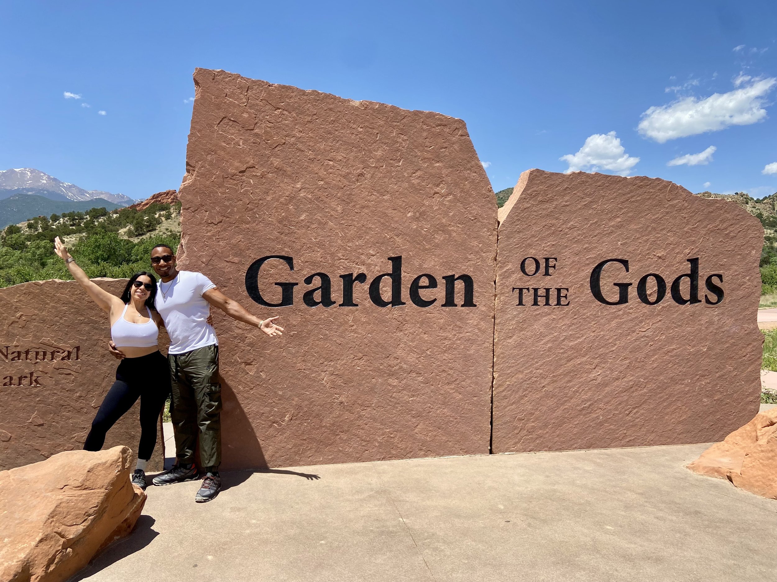 Two people standing in front of a large stone sign that reads "Garden of the Gods" in a sunny outdoor setting with mountains and trees in the background.