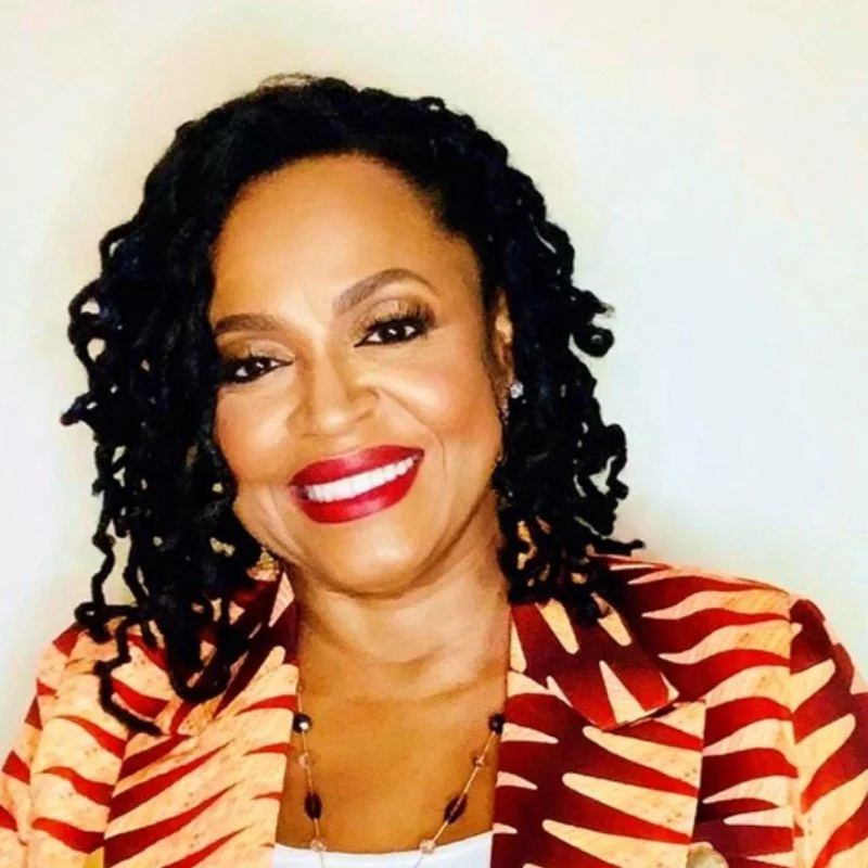A smiling woman with dark, curly hair, red lipstick, and earrings, wearing a red and beige patterned blazer.