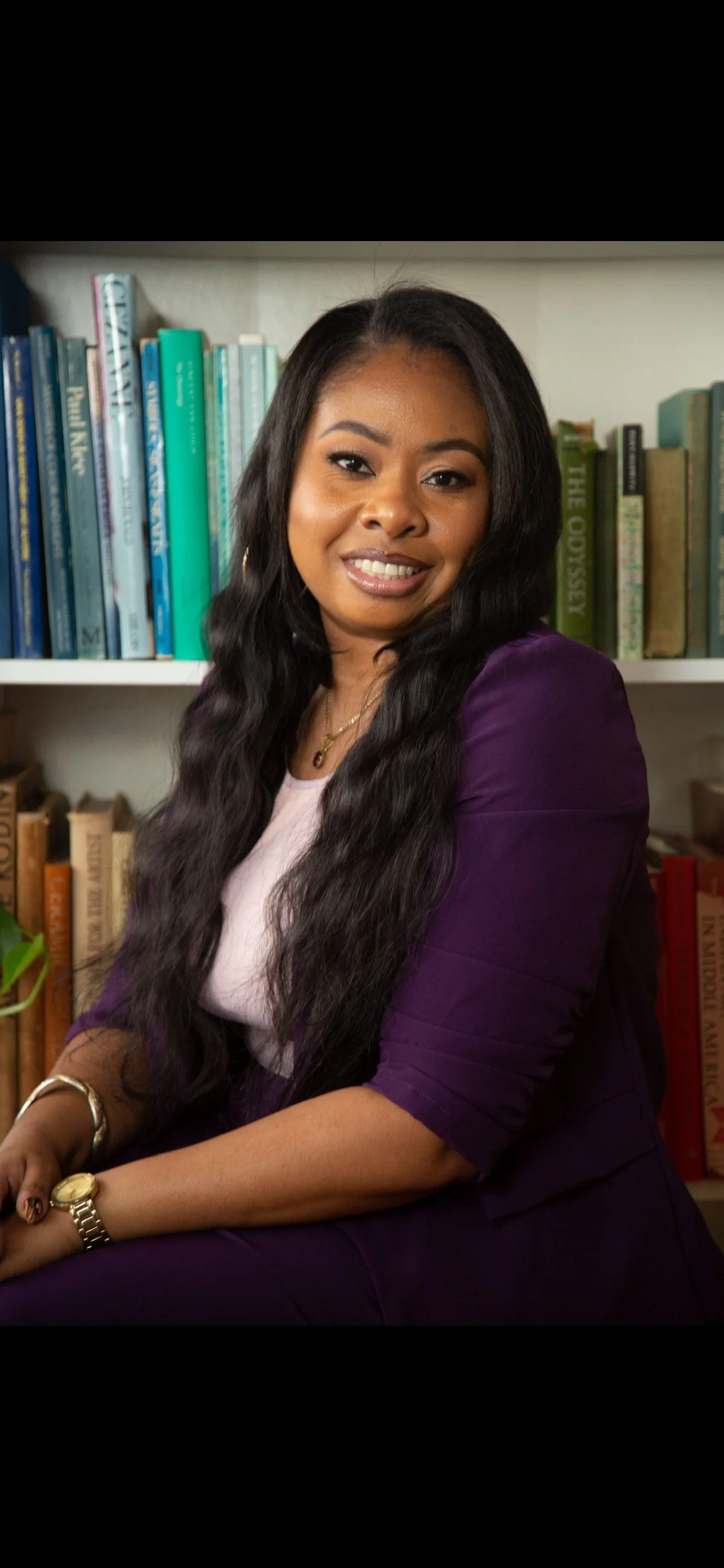 A woman with long black hair, wearing a purple blazer and a light pink top, sitting in front of bookshelves filled with books.