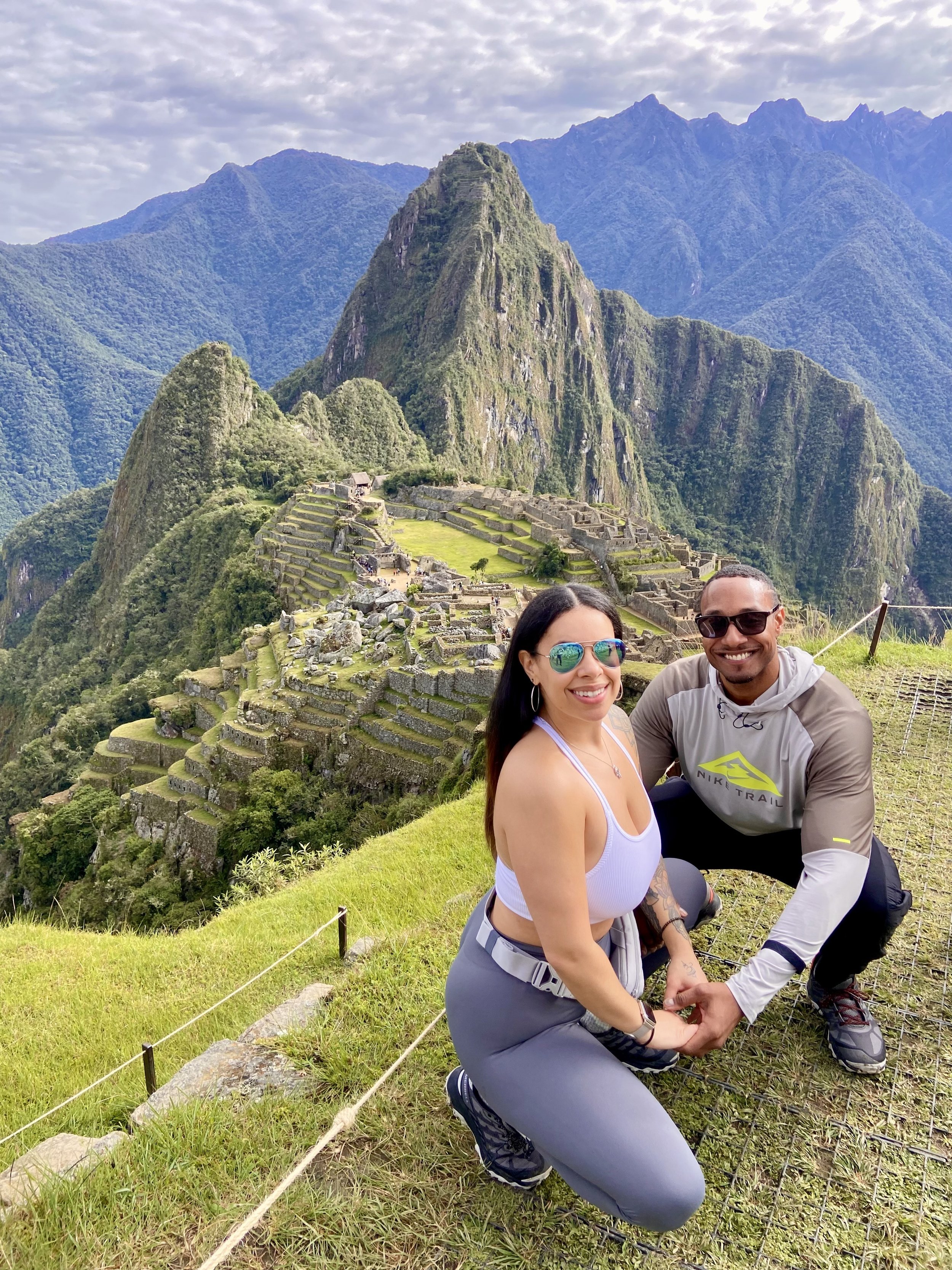 A smiling couple posing hand-in-hand with Machu Picchu ruins and mountains in the background.