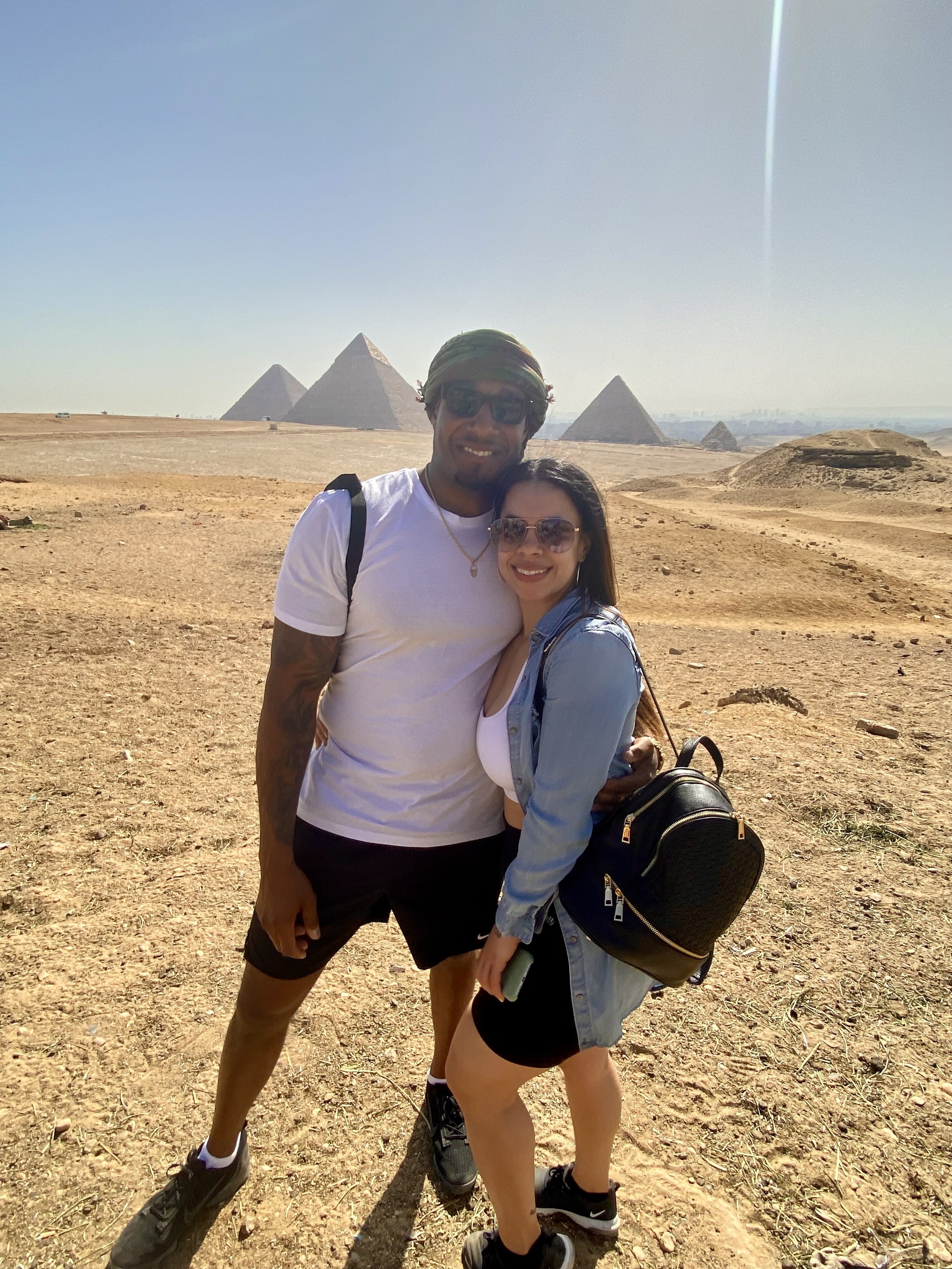 A smiling couple selfie on a desert landscape with the pyramids of Egypt in the background.