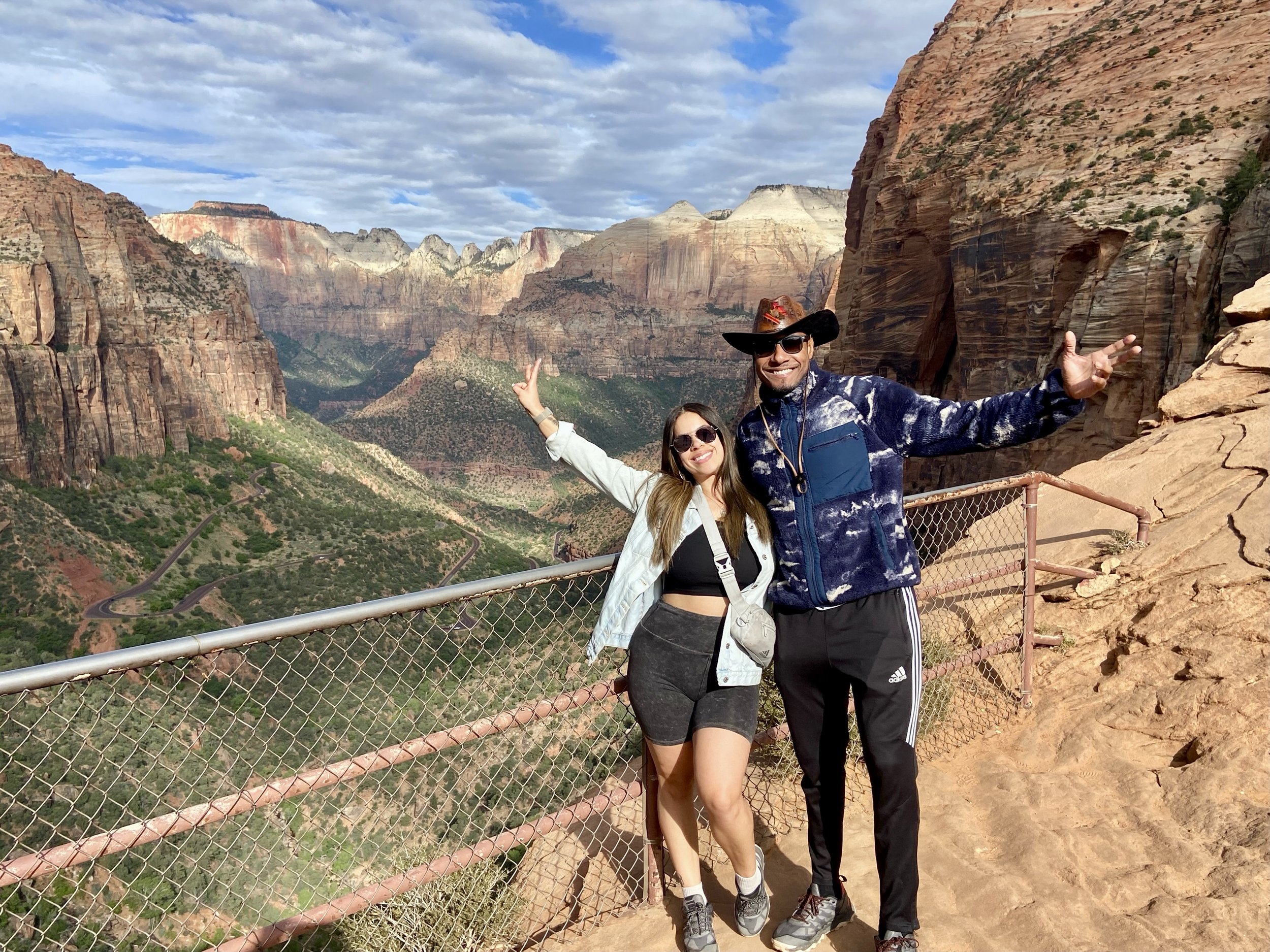 A smiling couple poses in front of the Grand Canyon, with cliffs and a winding road visible in the background.