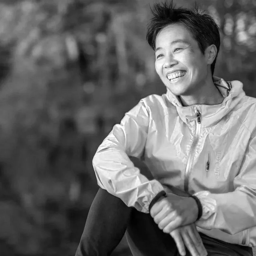 Eine lachende Frau sitzt draußen in der Natur in einem leichten, hellen Windjacke.