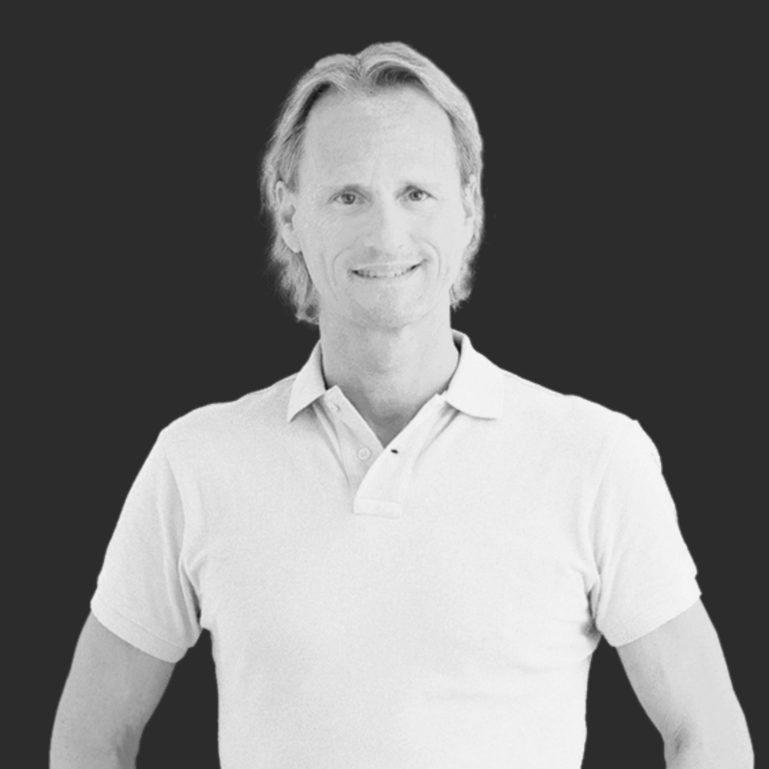 Schwarz-weiß Foto eines lachenden Mannes mit langen Haaren, trägt ein helles Polo-Shirt, vor dunklem Hintergrund.