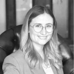 Porträt einer Frau mit langen Haaren und Brille, lächelnd, in einem dunklen Raum sitzend.