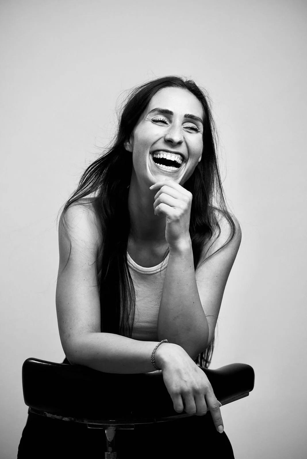 Schwarz-weiß Bild einer lachenden jungen Frau, die sich auf einem Stuhl abstützt, mit langem dunklem Haar, Tanktop und Armbändern.