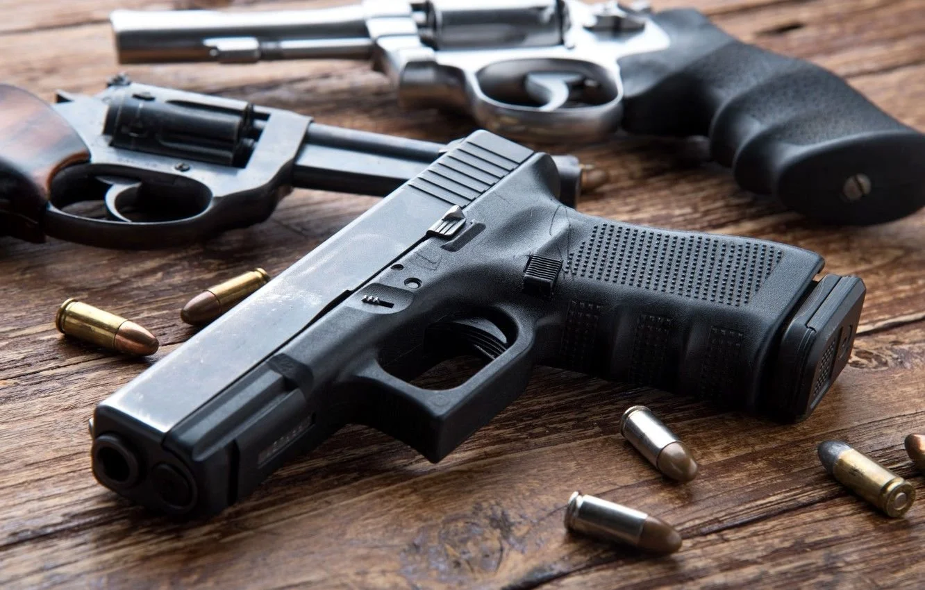 Three handguns with bullets scattered around on a wooden surface.