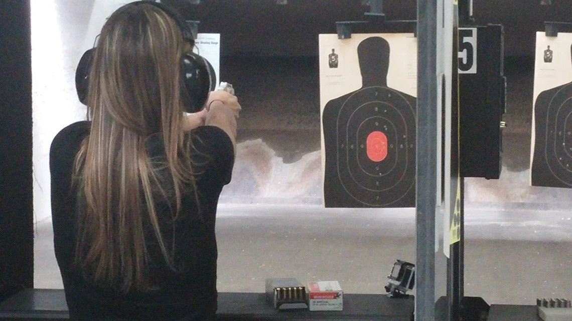 A woman with long hair and hearing protection aiming a handgun at a shooting range with target silhouettes.