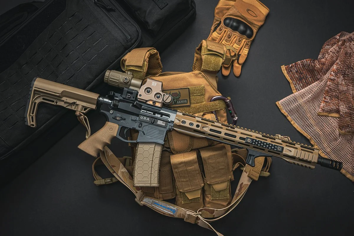 A tactical firearm with tan and black components, including a scope, mounted on tactical gear such as a camouflage glove, a tan vest with pouches, and a black carrying case, on a dark surface.