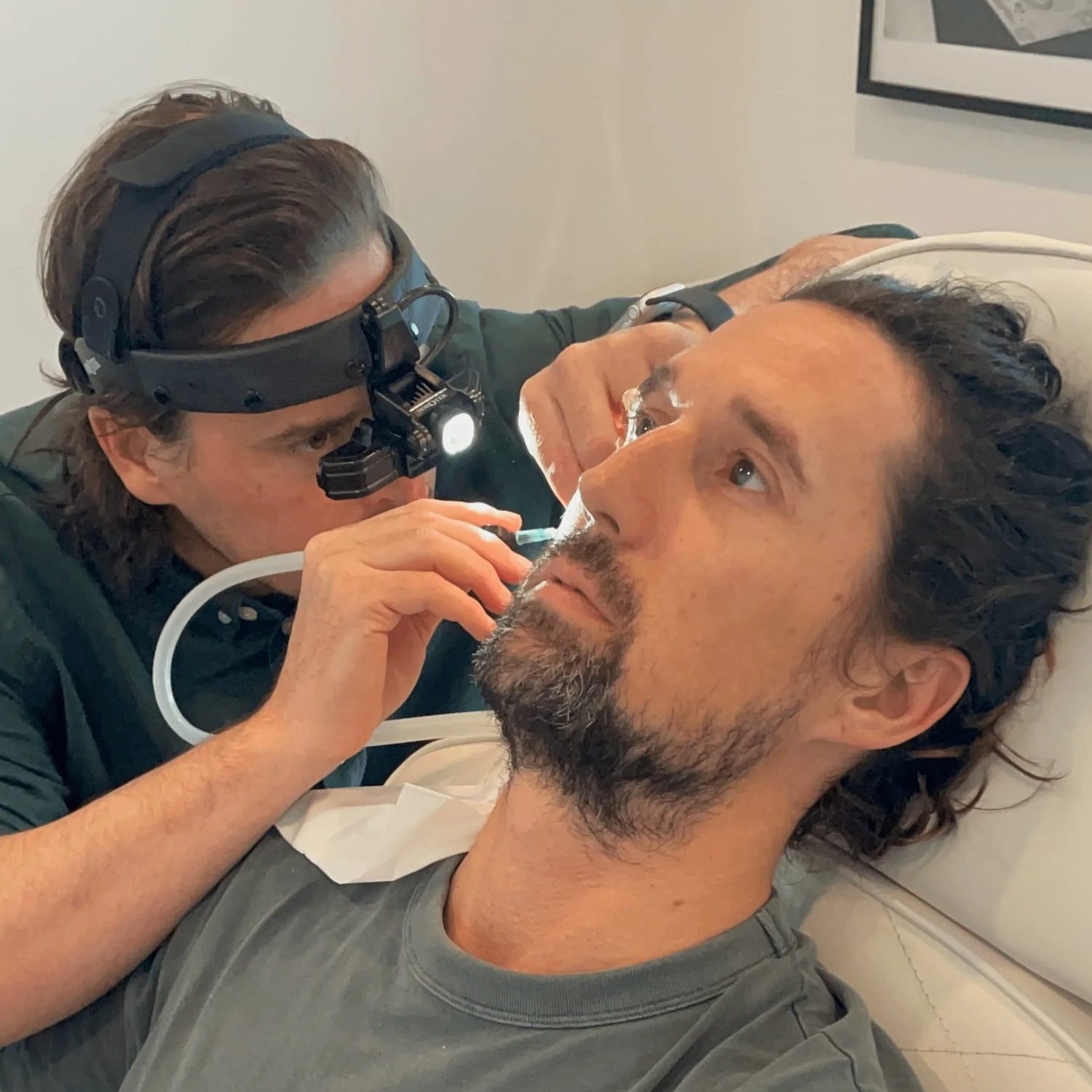 A healthcare professional examines an adult male patient lying on a medical examination table, looking up at the professional. The professional wears a headlamp and appears to be performing a medical procedure on the patient's face.
