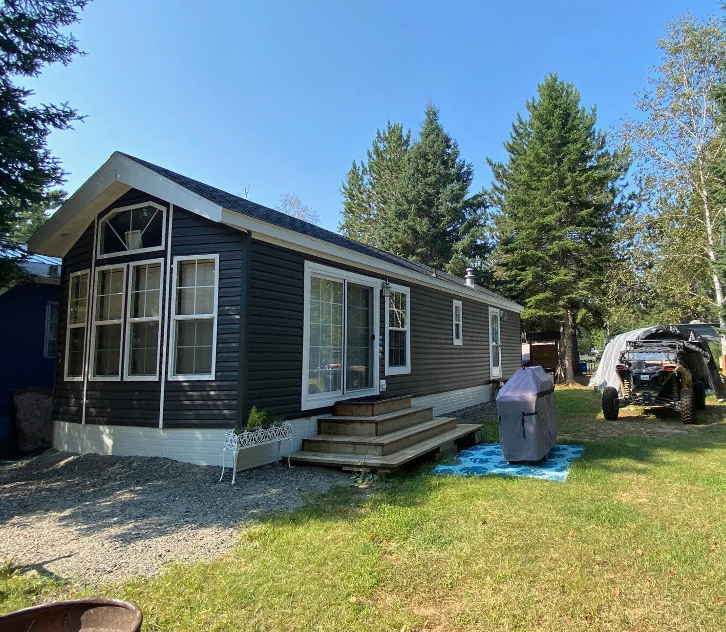 Mobile home with gray siding and white trim, surrounded by grass and trees, a small deck with steps, and a covered BBQ grill and all-terrain vehicle on the lawn.