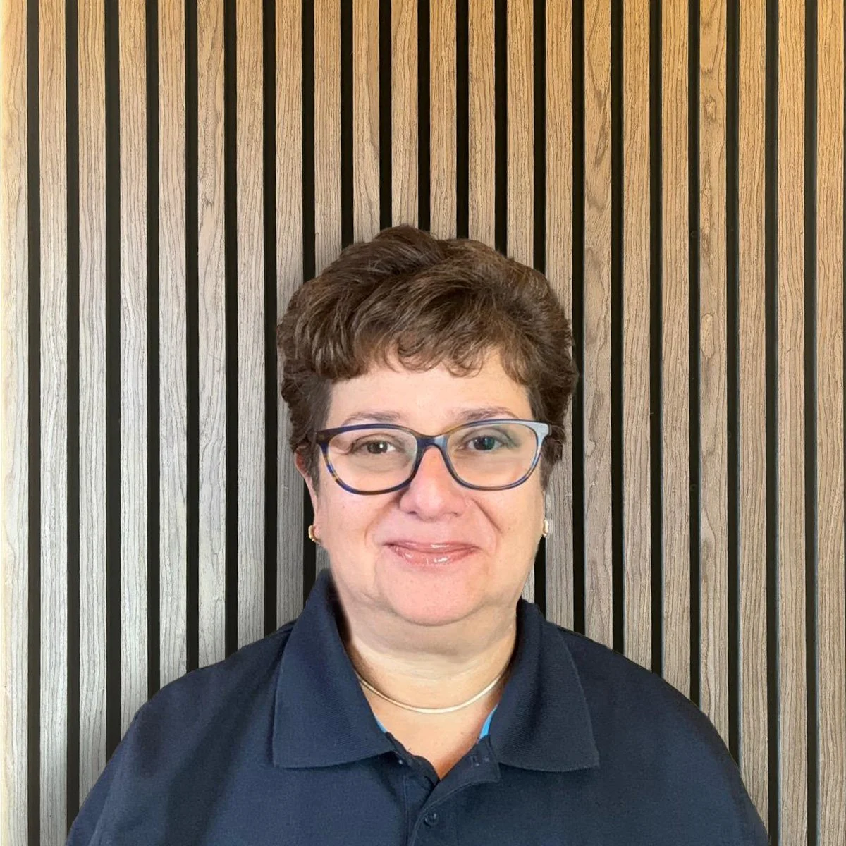 A woman with glasses and short curly hair, wearing a black polo shirt with the logo 'Elite' on it, smiling while sitting at a table.