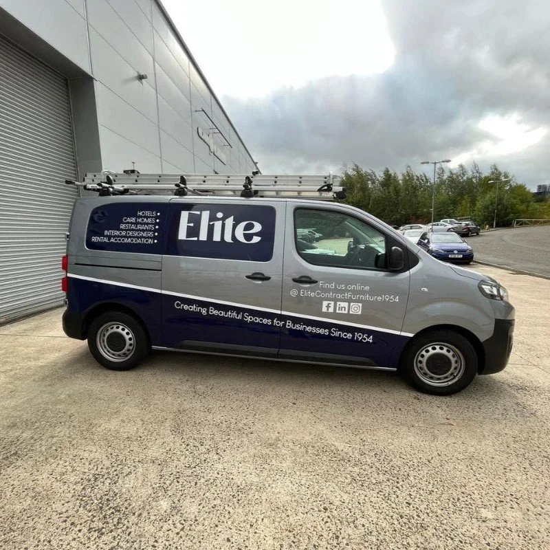 A commercial van parked outside a large building on a cloudy day. The van has branding for Elite, a business creating spaces for hotels, interior designers, and rental accommodations. The van also shows social media icons and the company's online presence.
