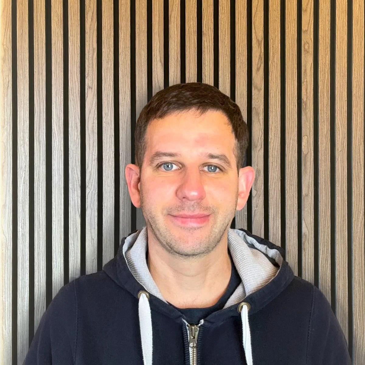 Portrait of a young man with short dark hair wearing a black zip-up jacket with an "elite" logo, standing in front of a colorful, patterned wall with botanical and animal motifs.