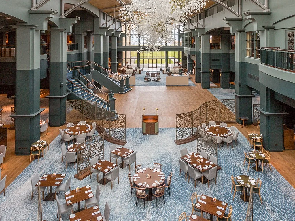 Interior view of a spacious hotel ballroom with round and rectangular tables set for dining, a dance floor, a staircase, and modern decor with large windows and a high ceiling decorated with hanging floral arrangements.
