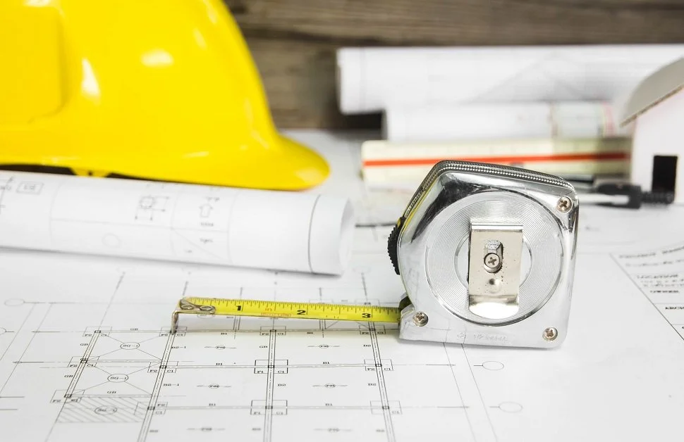 Construction plans, a yellow hard hat, and a metal tape measure on a work desk with blueprints.