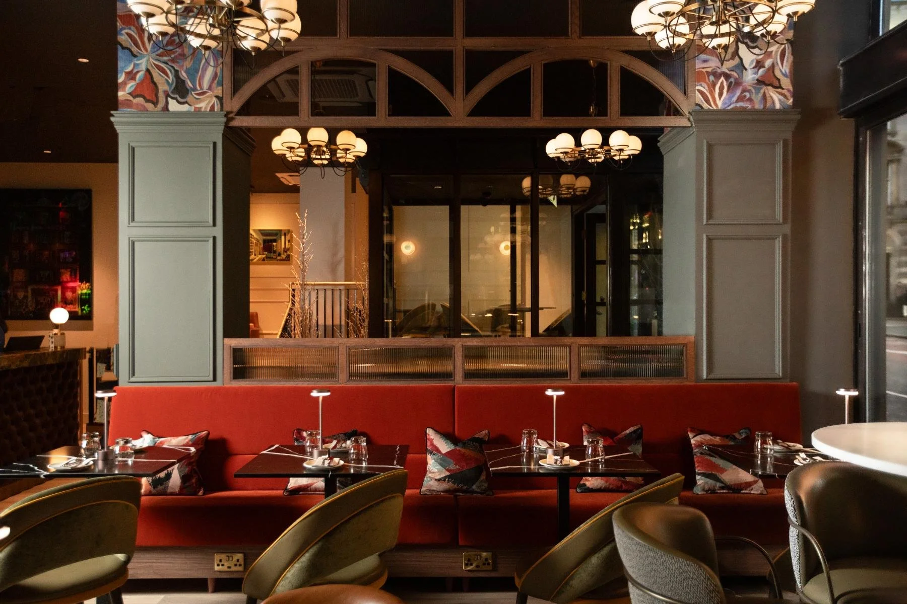Interior of a restaurant with red cushioned seating, pillows, tables set with glasses and cutlery, and ornate chandeliers hanging from the ceiling.