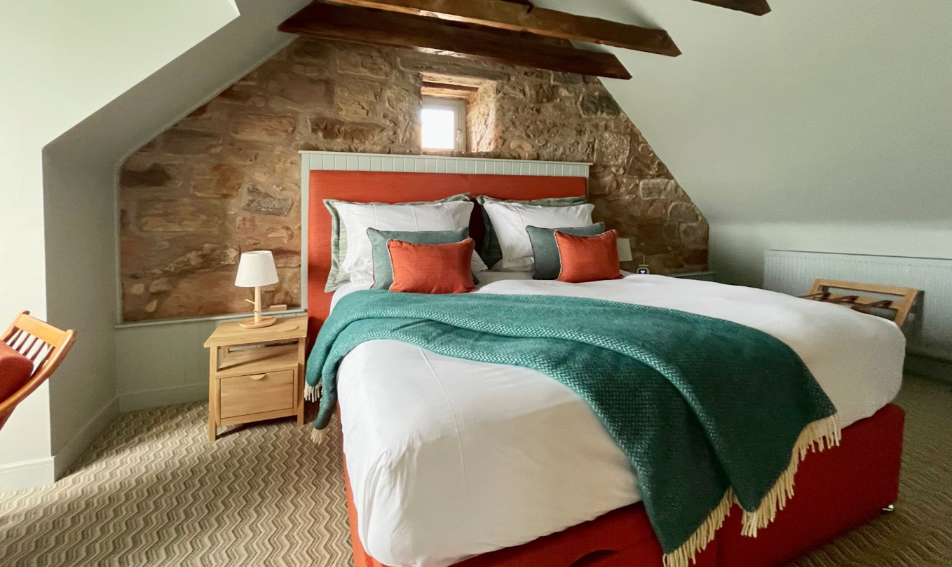 A cozy bedroom with a large bed, pillows, a green blanket, and a wooden bedside table with a lamp, set against a stone wall and a small window in the attic.