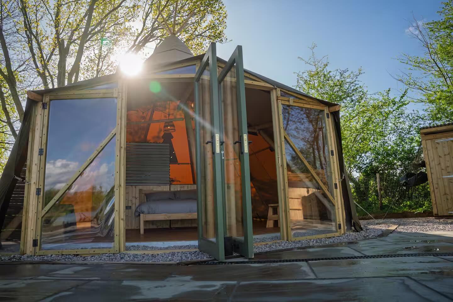 A small, transparent, geodesic-style building with wooden framing, set outdoors surrounded by trees, with sunlight shining behind it.