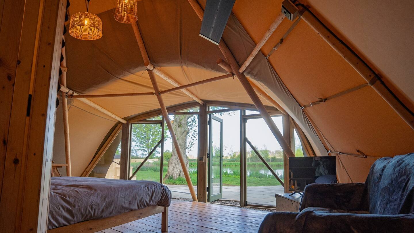Interior of a cozy glamping tent with a bed, a sofa, a TV, and large windows overlooking a green landscape with a tree and a pond.