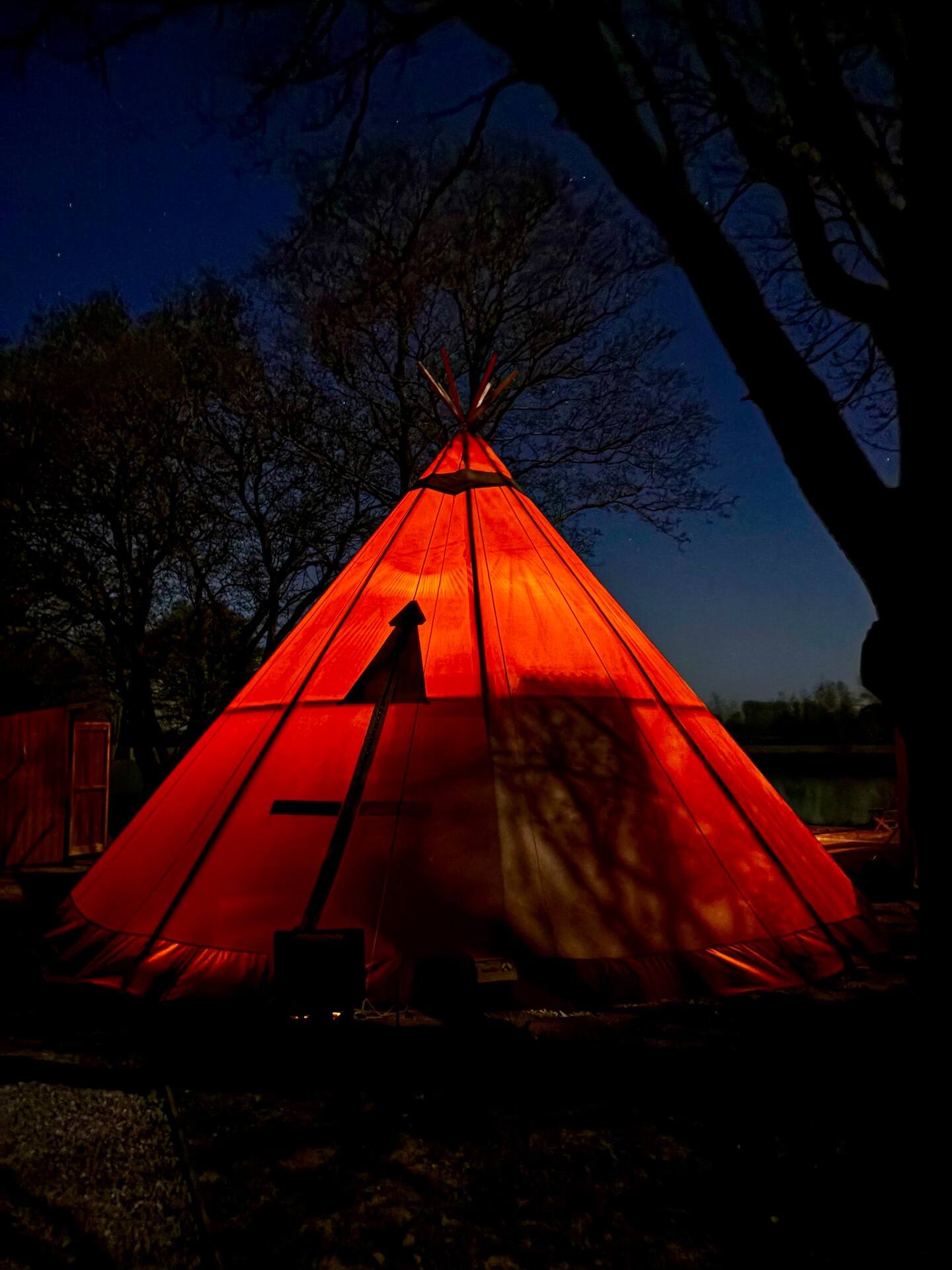 A large orange teepee tent set up outdoors at night with trees surrounding it, and a dark sky with some stars visible.