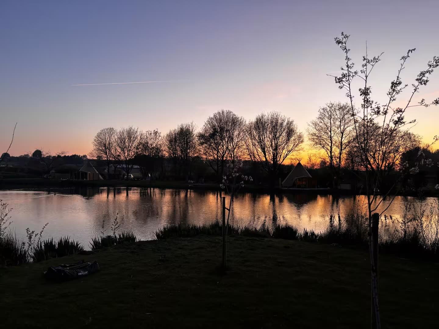 A peaceful scene at sunset with a lake reflecting the colorful sky, surrounded by bare trees and houses in the distance.