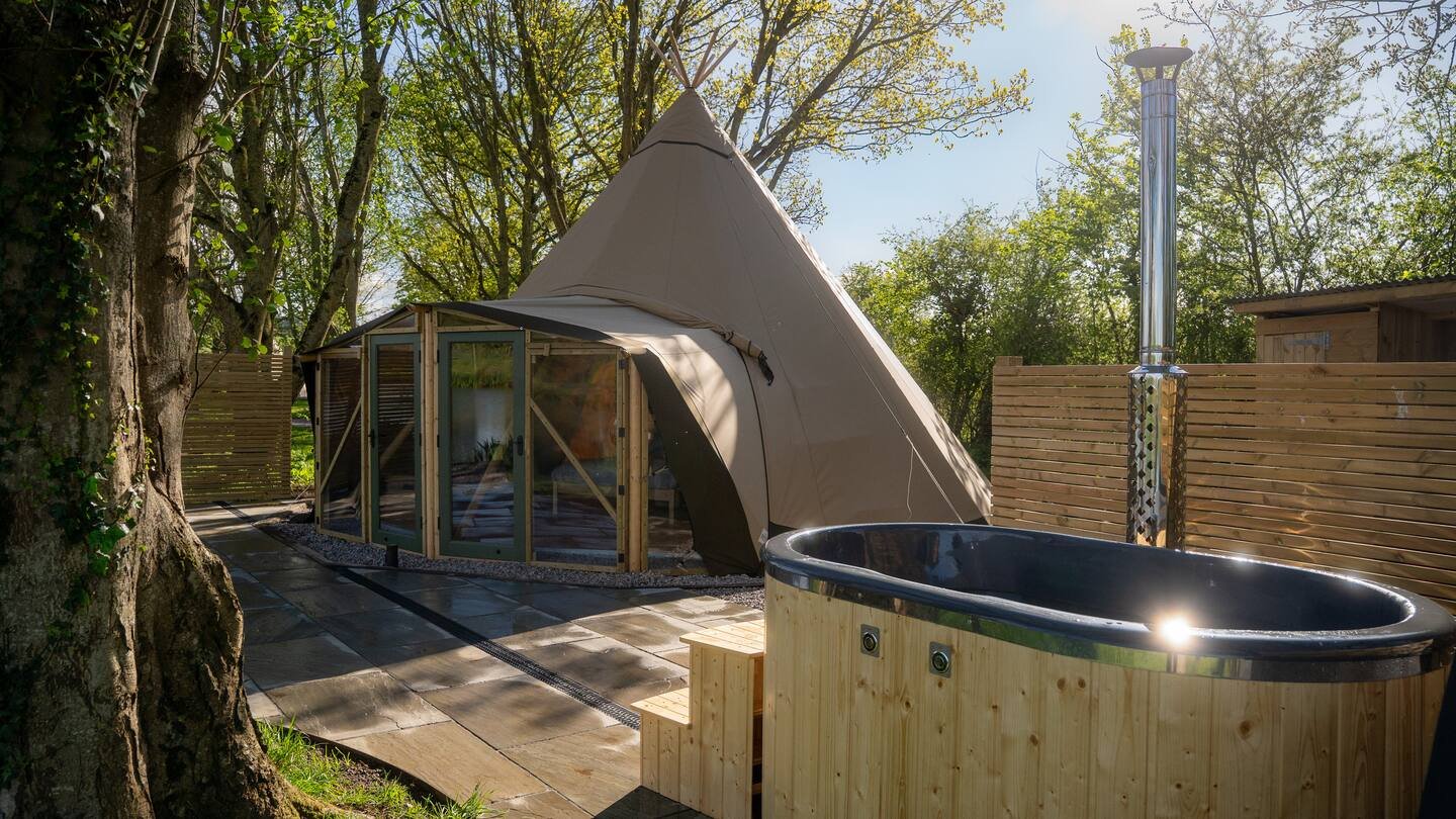 A backyard scene with a large beige teepee tent, a hot tub with a wooden exterior, a tall outdoor shower, and a wooden privacy fence. Trees and foliage are visible in the background, with sunlight filtering through the leaves.
