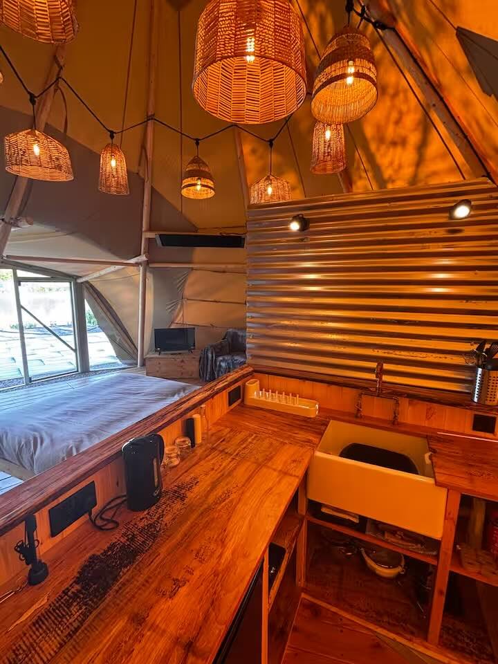 Cozy interior of a boutique hotel tent with hanging wicker lamps, a wooden desk with a black telephone, a white sink, and a bed with a window view of the outdoors.