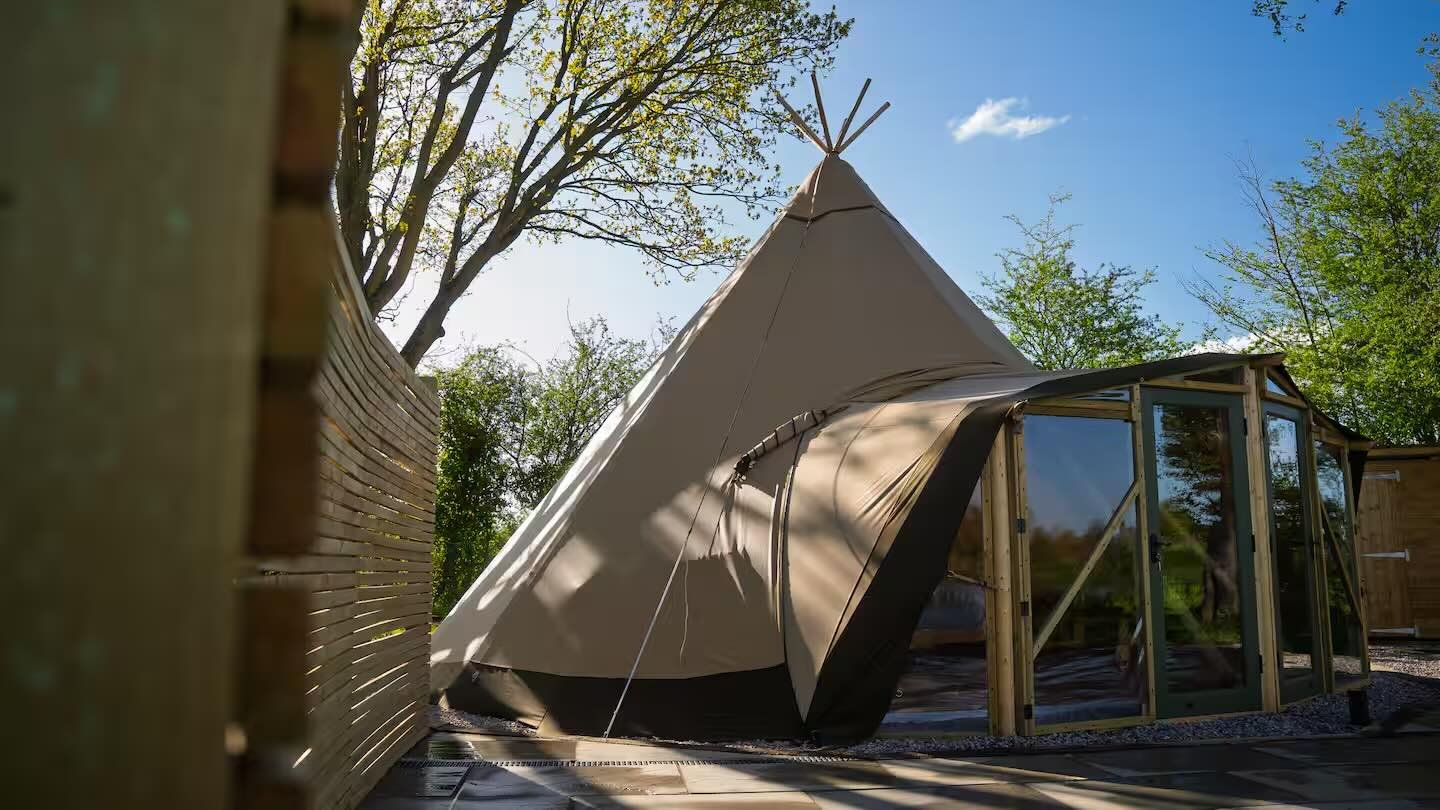 A large beige canvas tent with a teepee shape next to a modern structure with glass doors, set outdoors with trees and blue sky in the background.