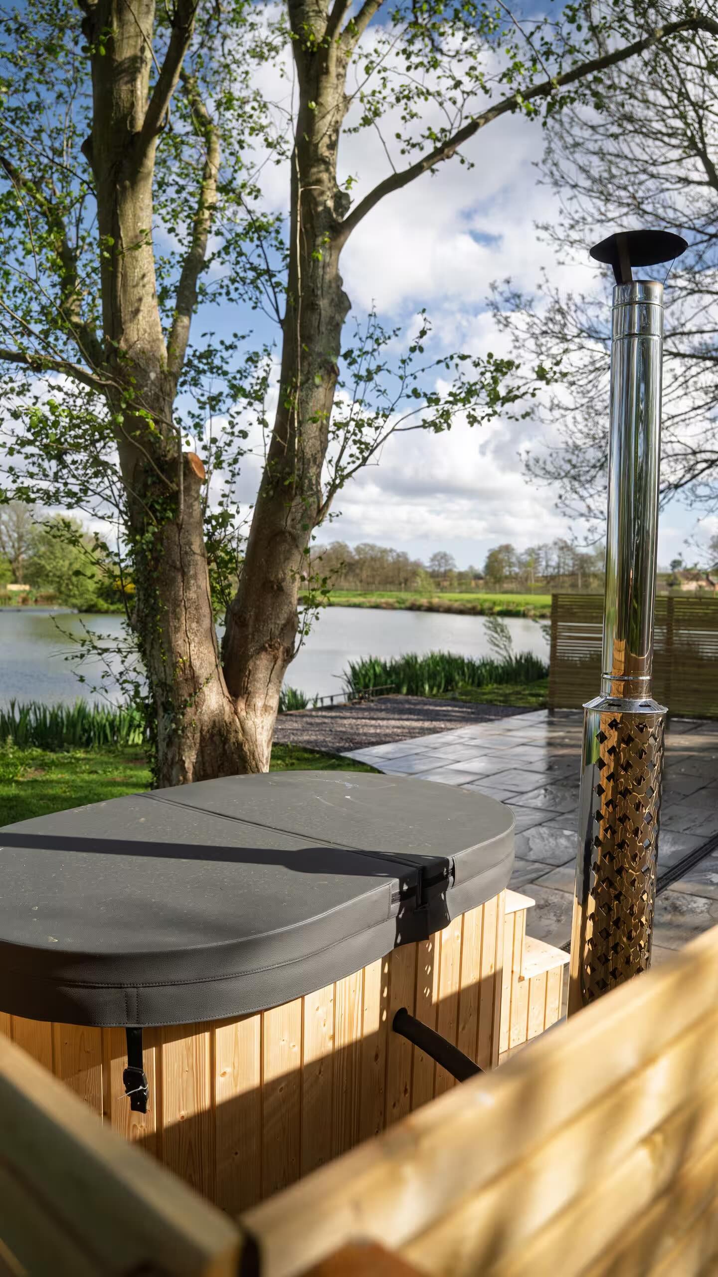 Outdoor hot tub with a cover next to a tree, overlooking a lake with greenery and a cloudy sky.