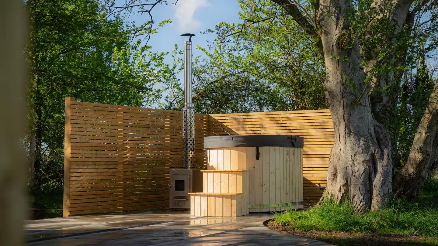 An outdoor wood-fired hot tub with a black cover, enclosed by a wooden fence, set next to a large tree with green leaves, under a clear blue sky.