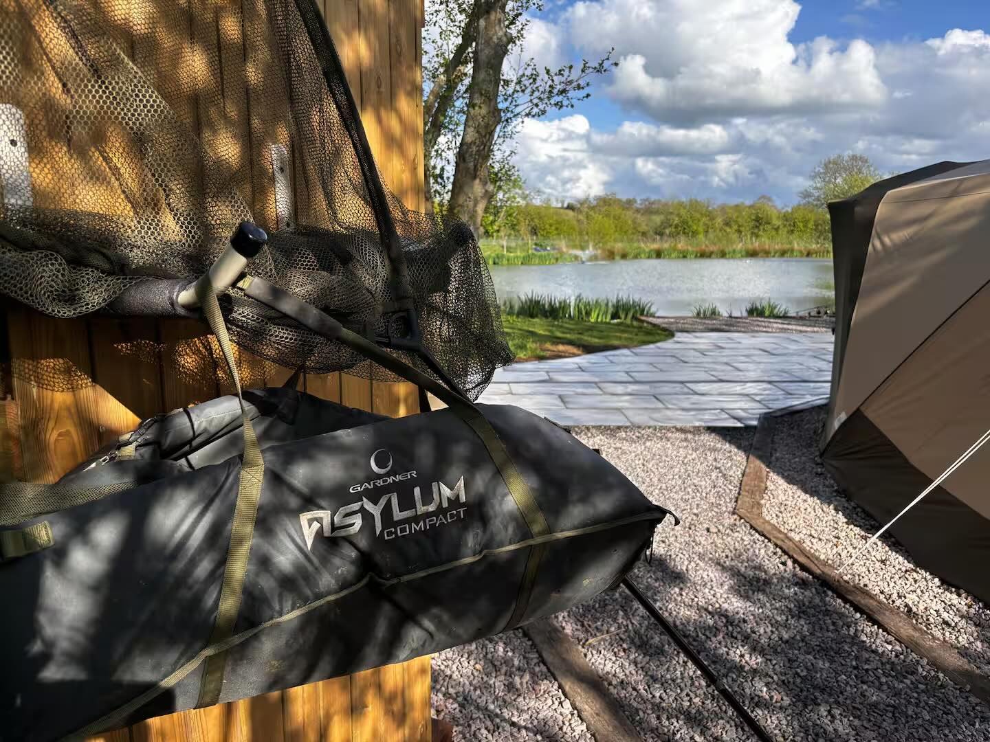 A portable fishing rod and tackle bag labeled "GARDNER ASYLUM COMPACT" rest against a wooden wall in an outdoor fishing spot. In the background, a lake surrounded by green trees under a partly cloudy sky is visible, with a gravel pathway and a tent n