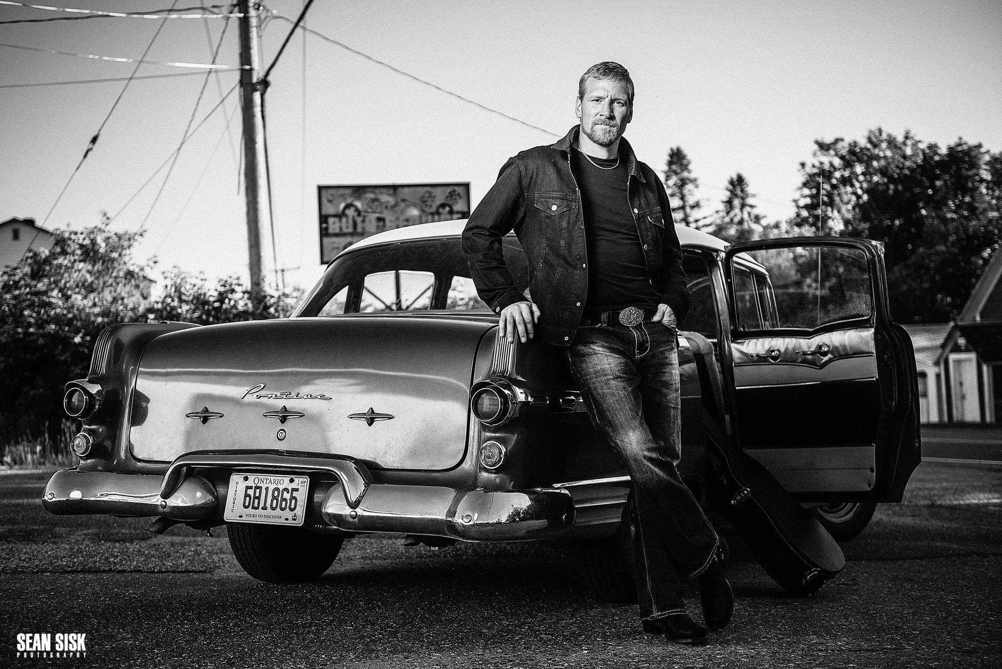 Danny Sylvester posing beside a vintage Pontiac in a black and white promotional photo