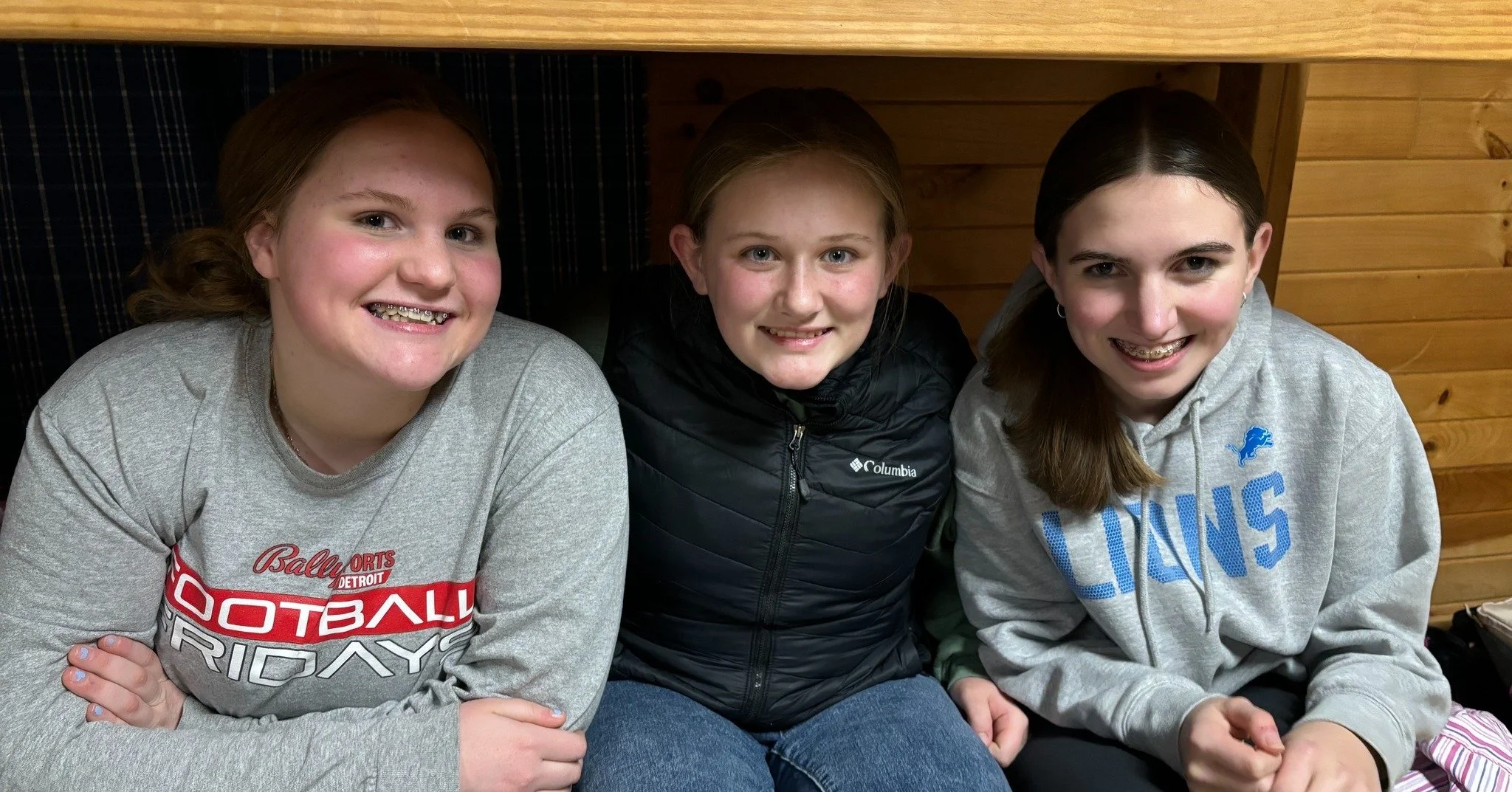 Three girls sitting closely together, smiling at the camera, in a room with wooden walls.
