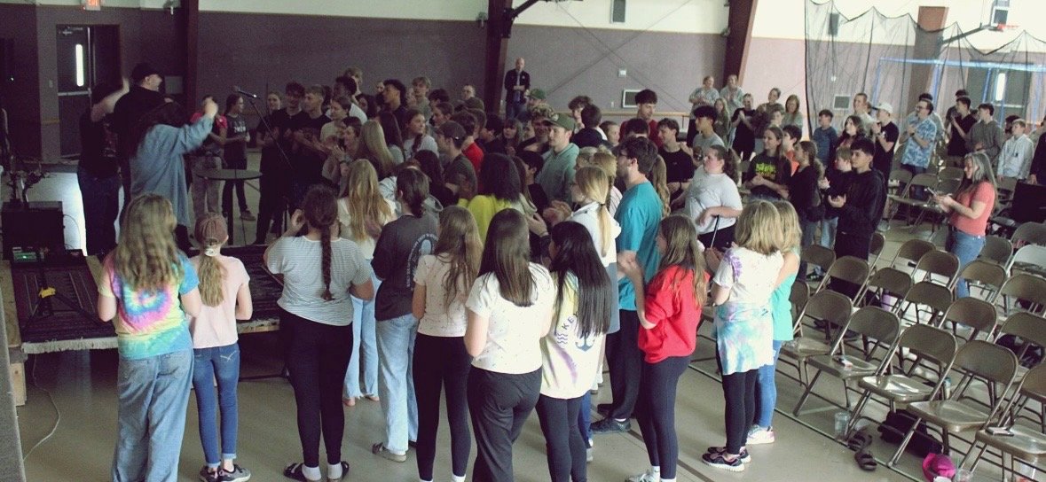 A group of children and teenagers gathered in a room, some standing in line or sitting, with a few adults present. The room has chairs and musical instruments, suggesting a school or community event.