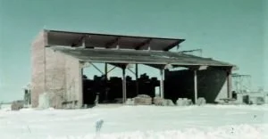 A partially constructed concrete building with a metal roof, situated in a barren, dusty landscape under a teal sky.