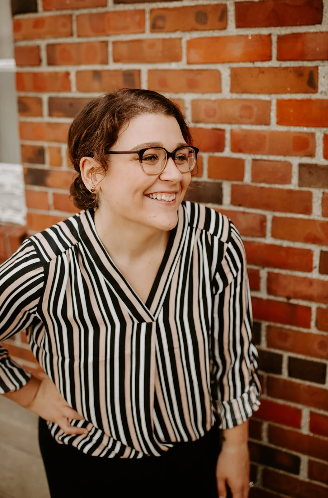 A woman with glasses and short brown hair, smiling and standing with her hand on her hip in front of a red brick wall.