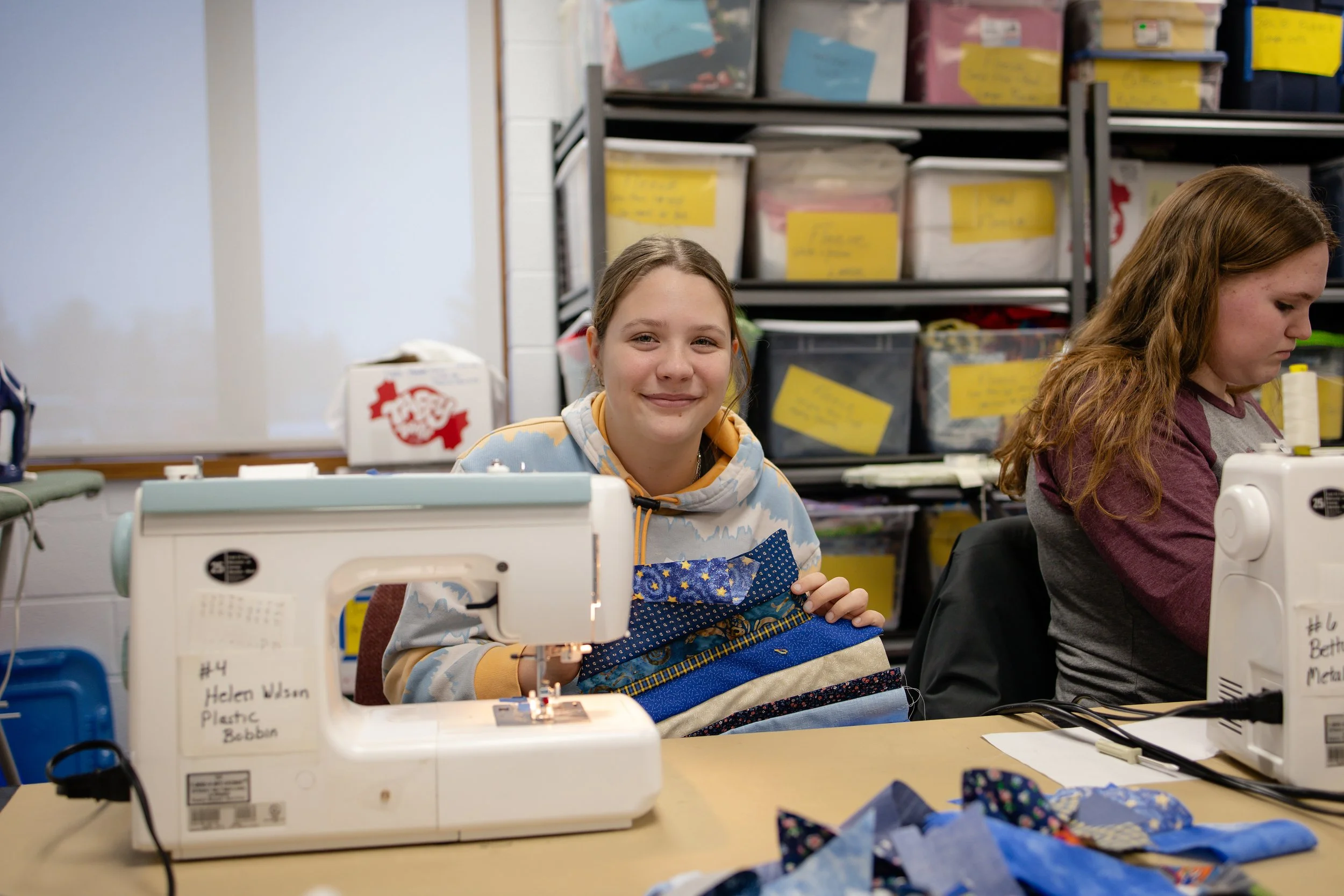 Two girls sitting at a sewing table working on fabric projects in a classroom. The girl on the left is smiling and holding a partially sewn piece of fabric. The girl on the right is focused on her sewing machine. Shelves with storage boxes are visibl