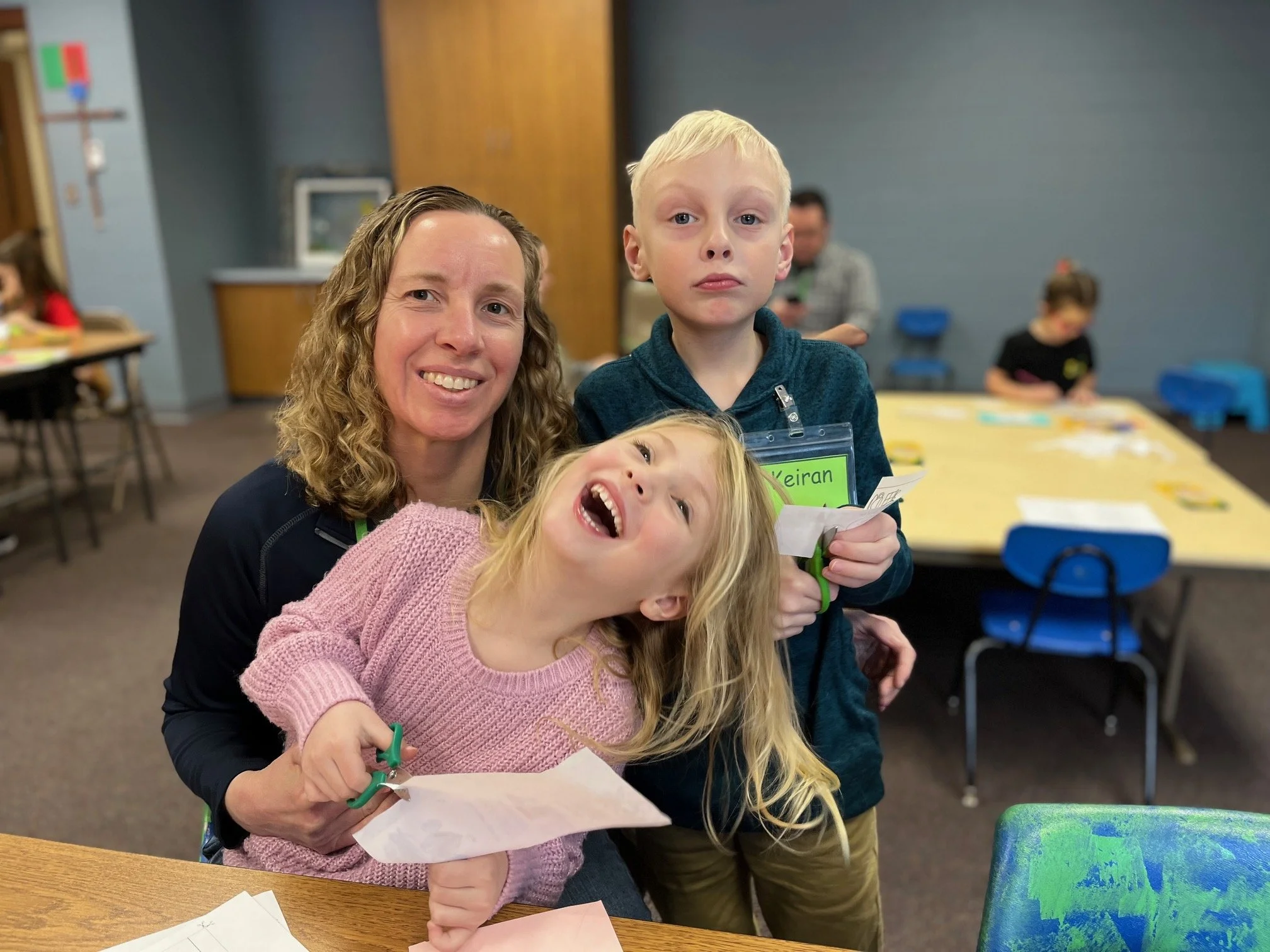 A woman with curly hair smiling while holding a young girl with blonde hair and a pink sweater, both standing behind a boy in a blue hoodie with a name tag, in a room with blue walls and children engaging in activities at tables.