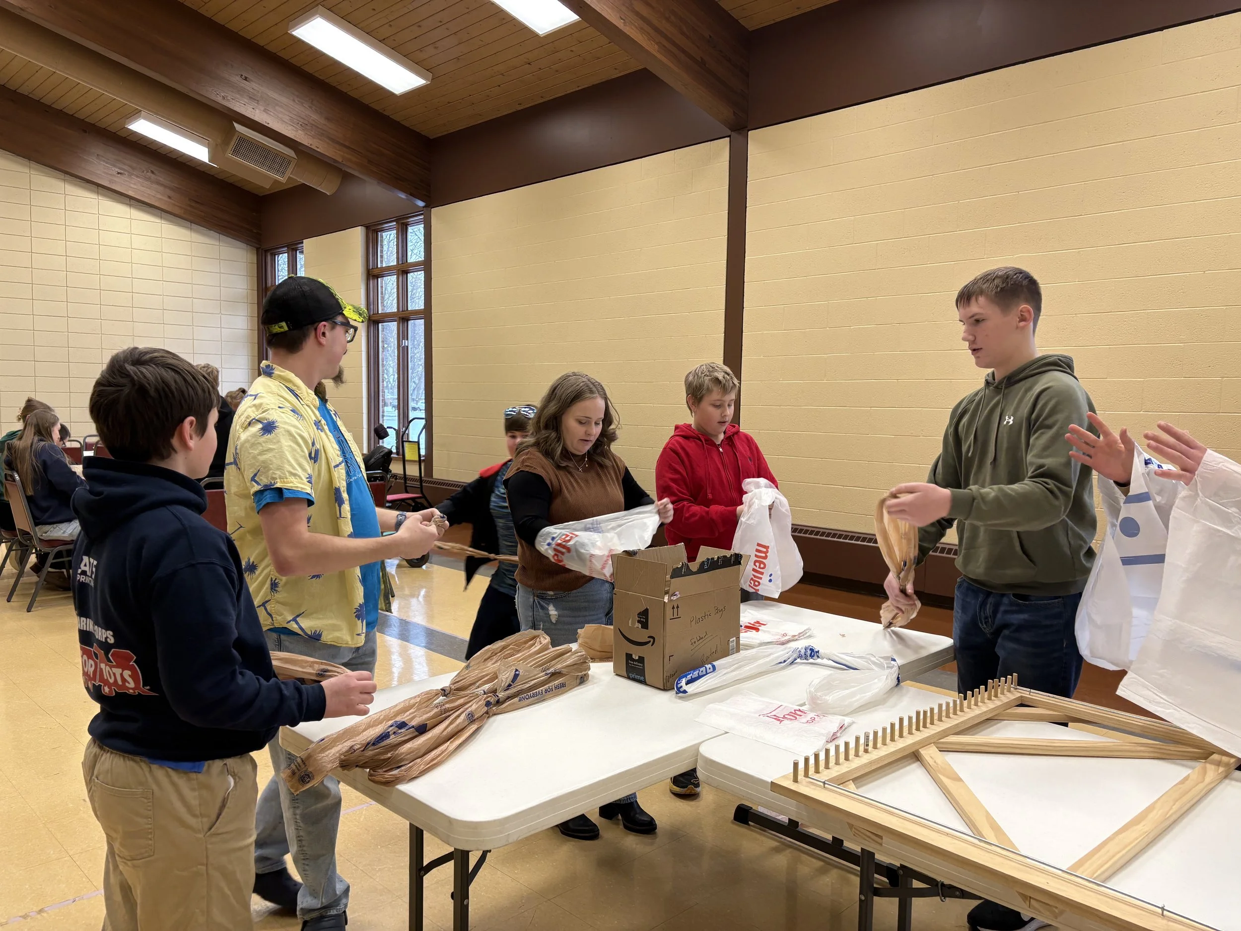 Group of children and adults gathered around a table set up for a craft or activity event inside a room with beige walls and wooden ceiling with large windows in the background.