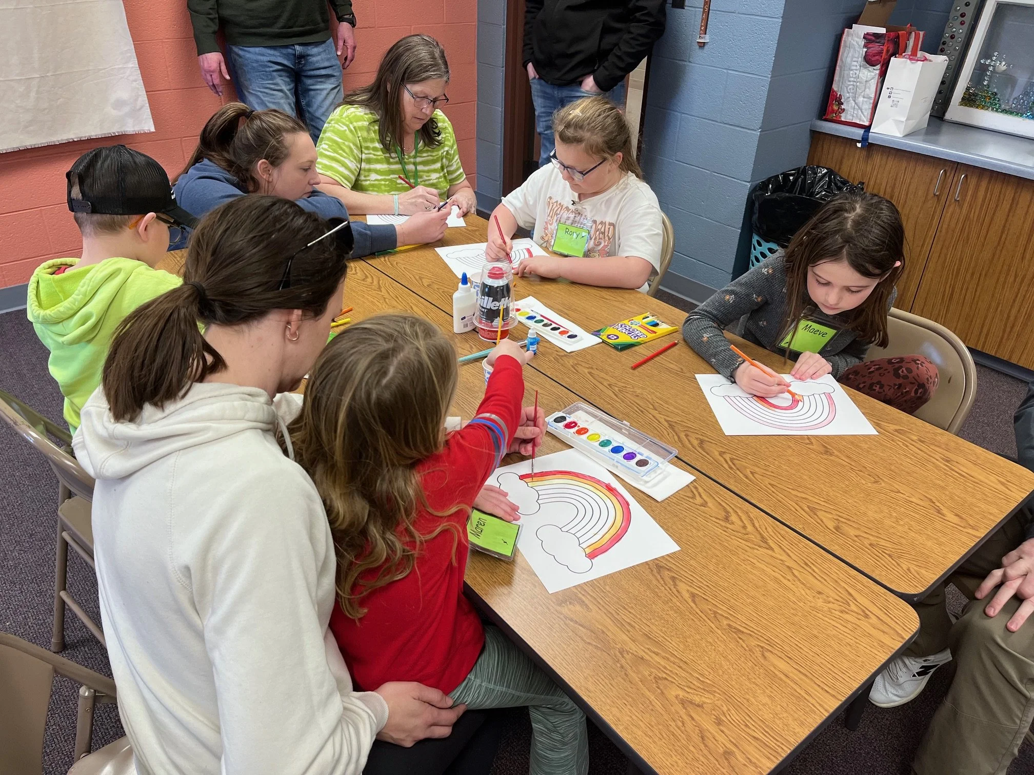 Several children and a woman are sitting around a wooden table, coloring rainbow and cloud pictures with watercolor paints and colored pencils. The children are focused on their artwork, and some are wearing name tags. The room has blue and orange wa