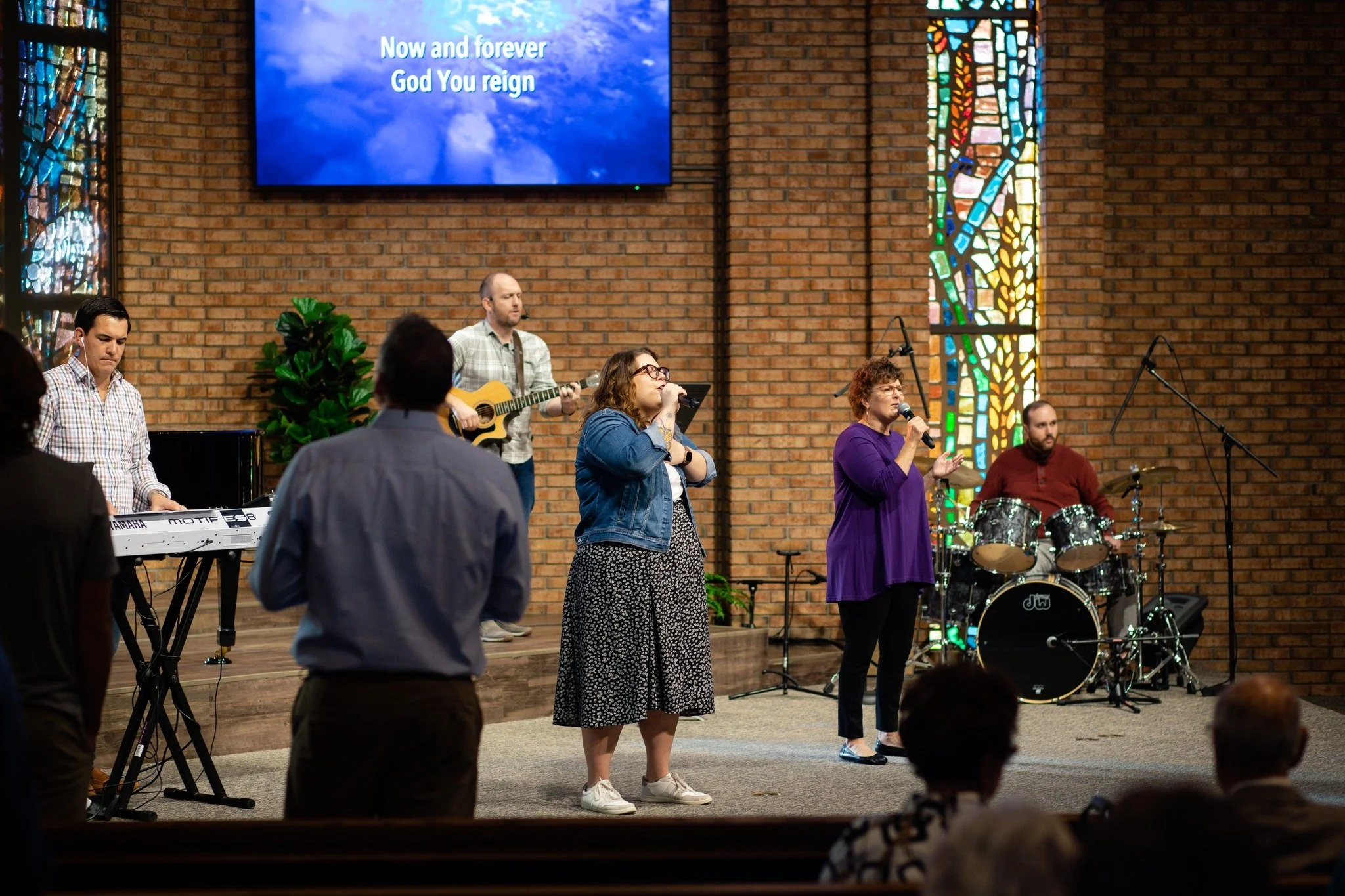People singing and playing music in a church service. Two women are holding microphones, one man is playing guitar, another man is playing drums, and a person is standing at a keyboard. There are stained glass windows and a digital screen with song lyrics in the background.