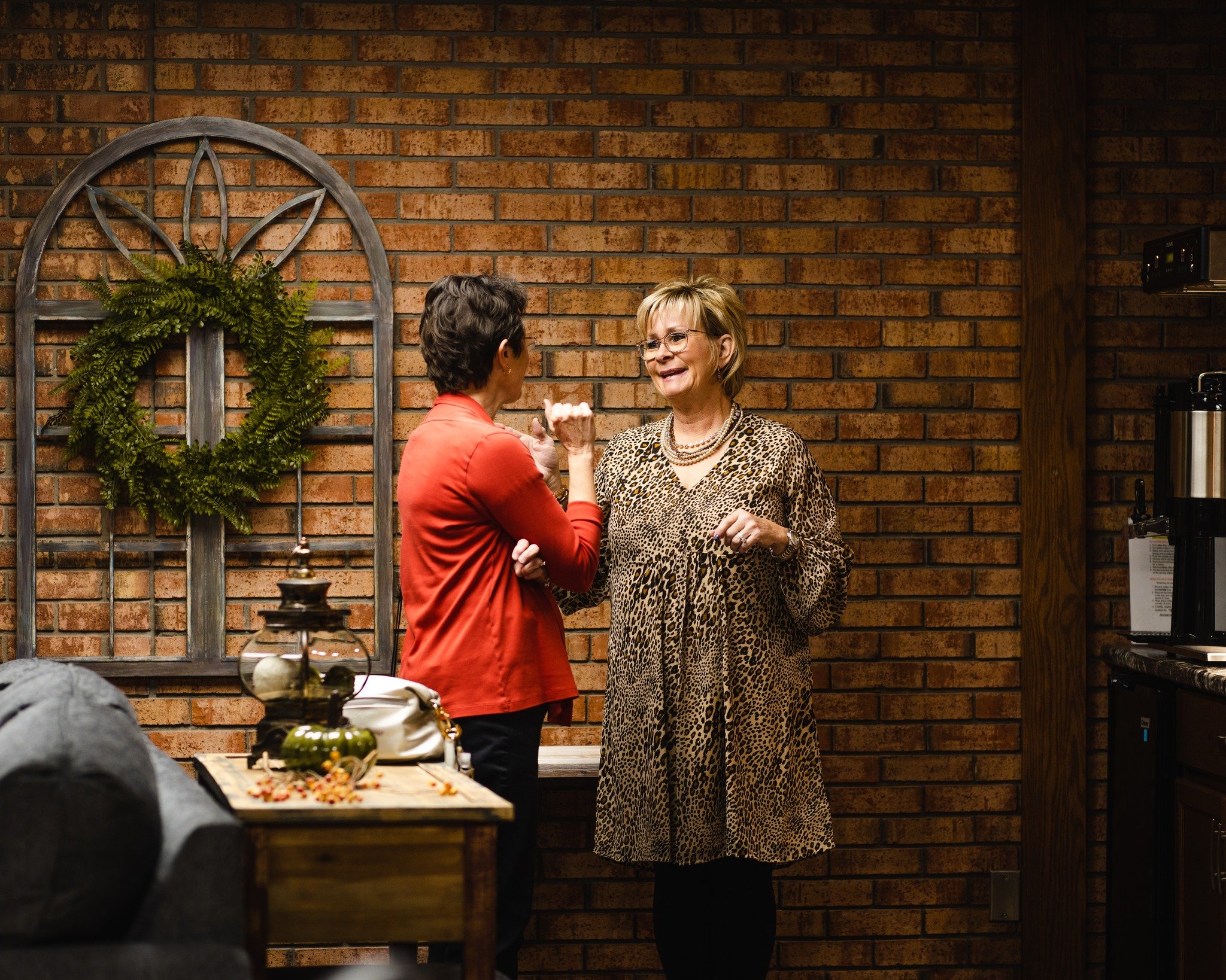 Two women are talking and smiling in front of a brick wall in an indoor setting. One woman is wearing glasses and a leopard print dress, while the other is dressed in a red blazer.