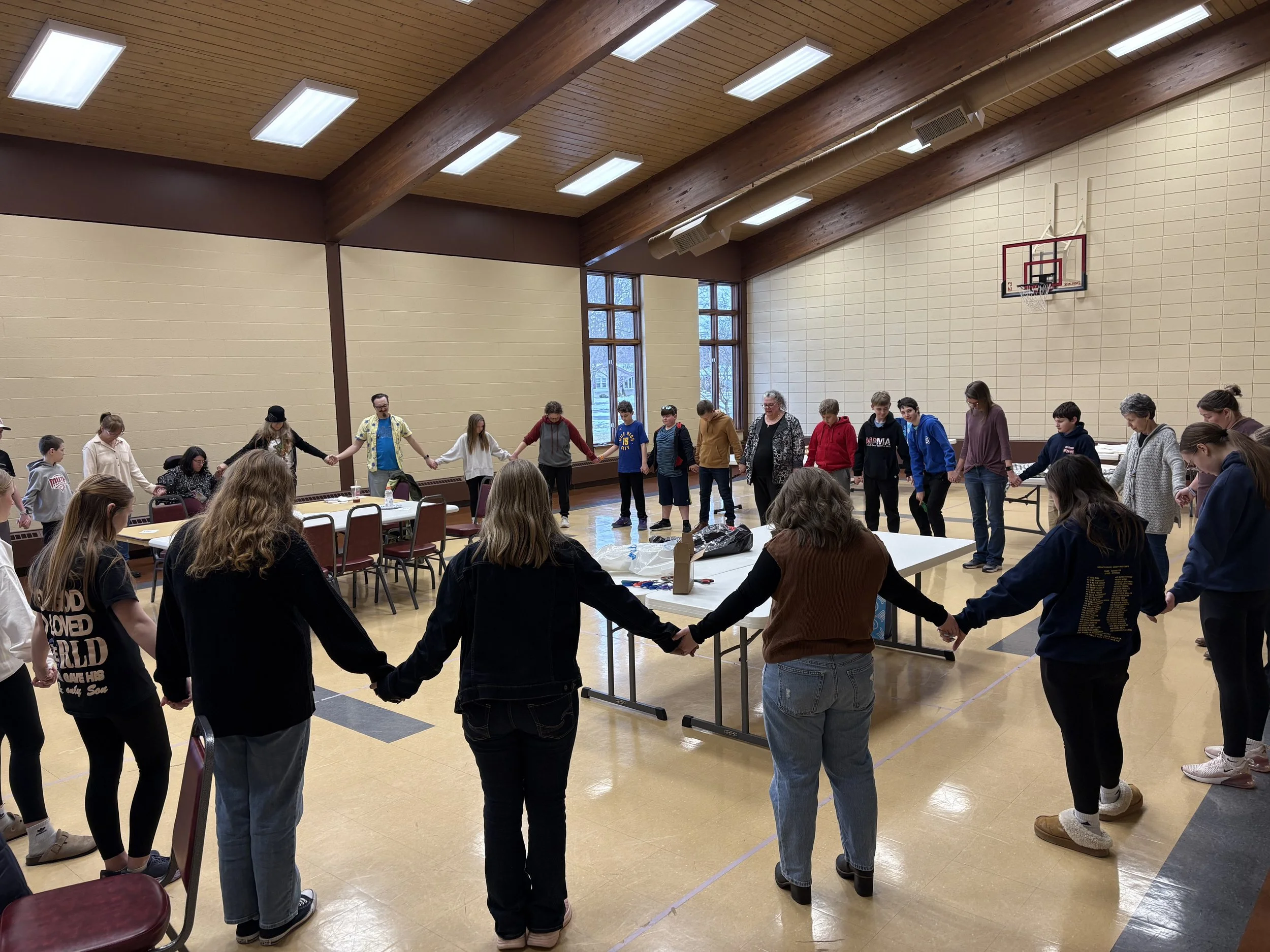 Group of people holding hands in a circle inside a gymnasium with a basketball hoop on the wall.