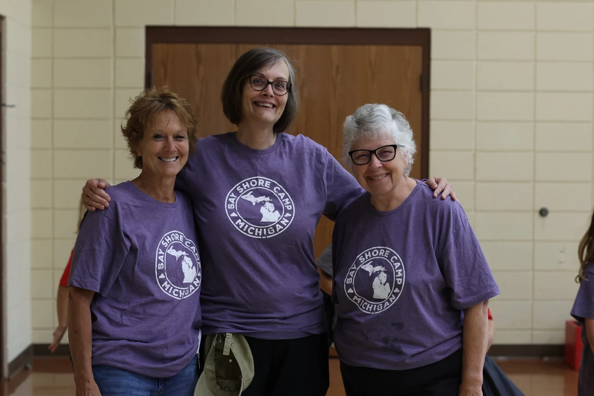 Three women smiling and standing together in matching purple t-shirts with 'Bay Shore Camp Michigan' logo, indoors, in front of a beige tiled wall.