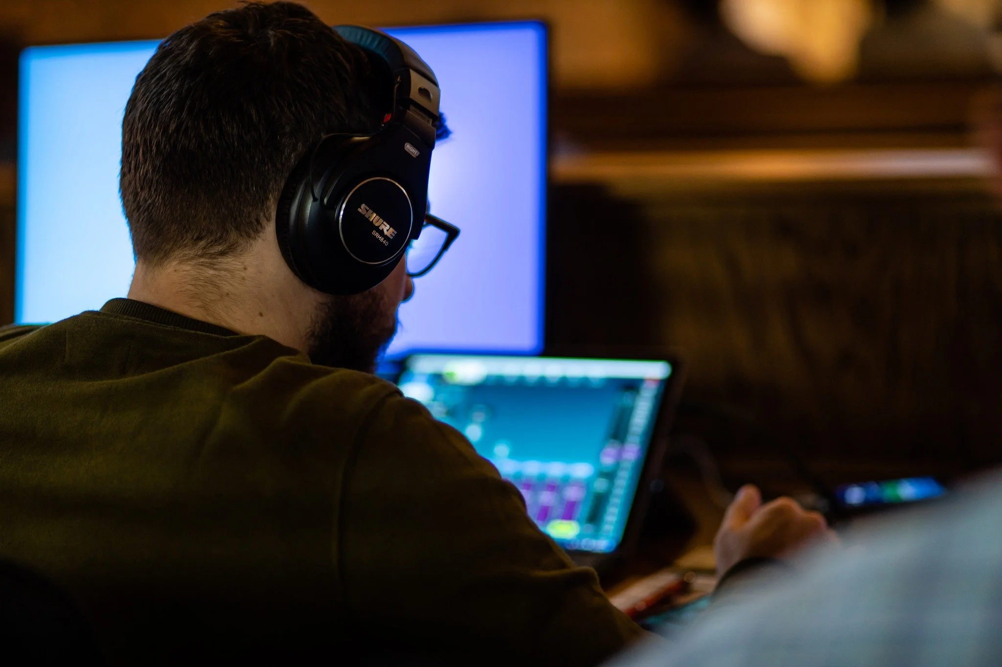 A person wearing glasses and a large black headphone with the brand name Shure, sitting in front of a computer monitor, with a lower left view of their head and back, indoors with warm lighting.