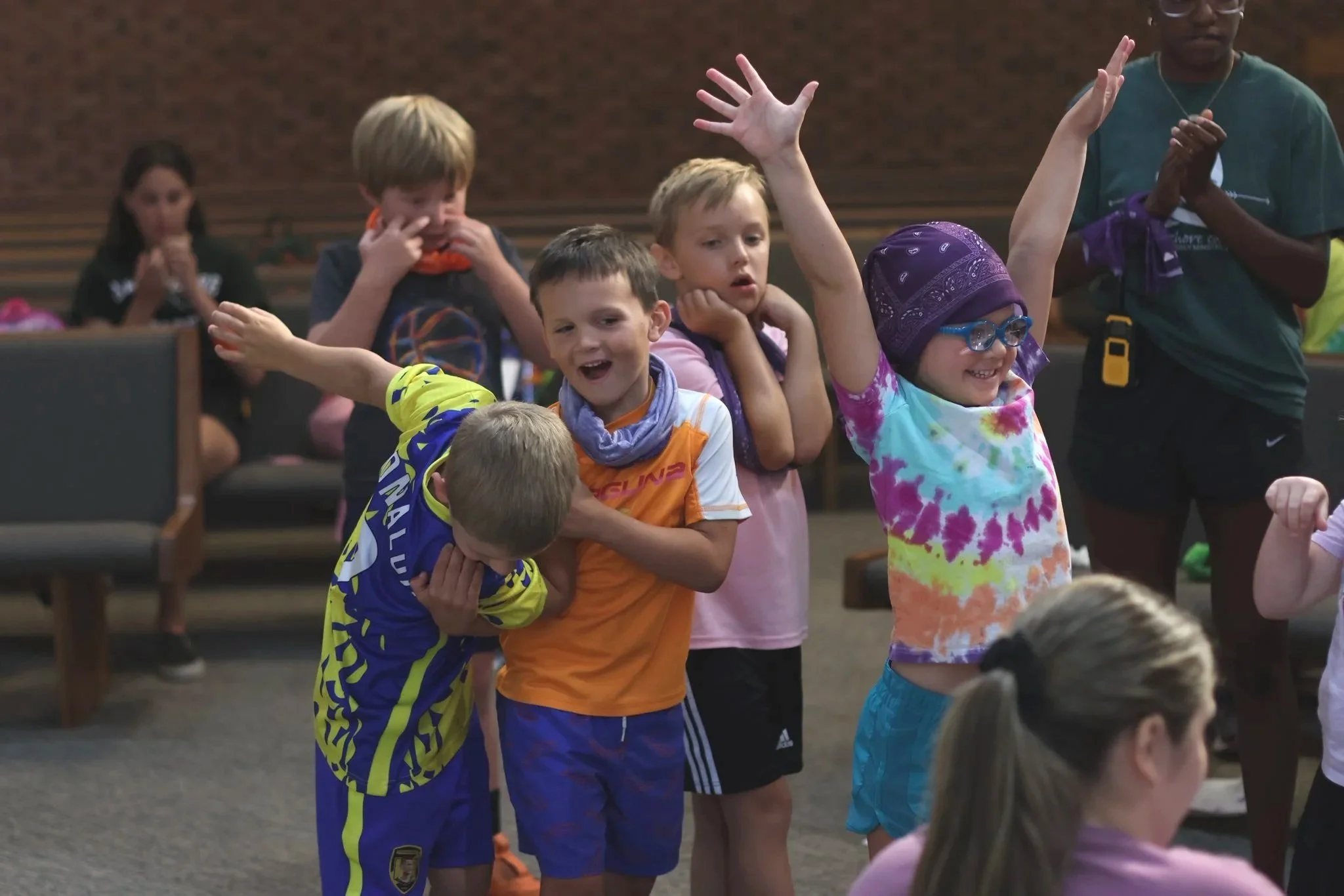 Group of children in a room, some with hands raised, participating in an activity. One girl with glasses and a purple bandana smiling, others showing various expressions of surprise or curiosity.