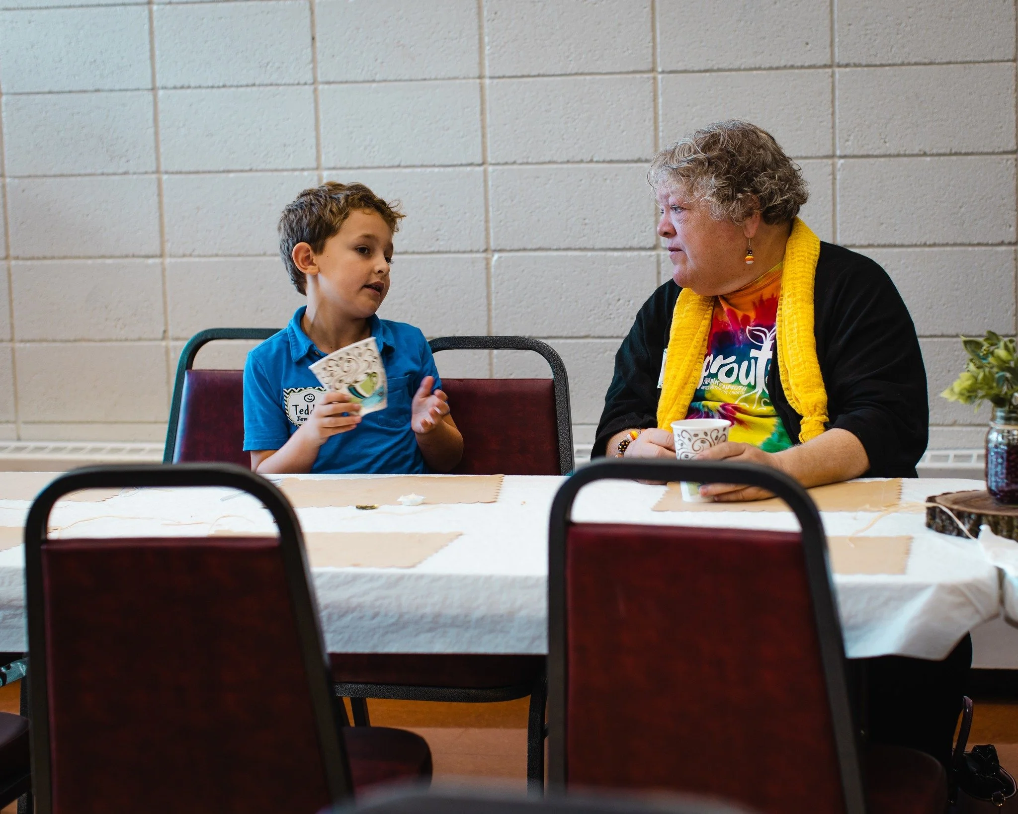 A boy and an older woman sitting at a table, engaged in conversation, with a plain brick wall in the background.