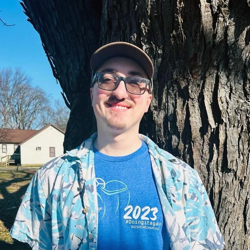 A young man with glasses and facial hair smiling, wearing a blue shirt and a patterned overshirt, standing next to a large tree on a sunny day.